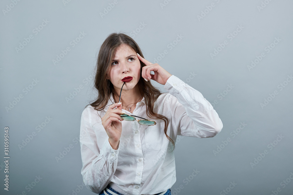 Attractive young brunette woman over isolated gray background wearing casual white shirt, holding fashion sunglasses, keeping finger on head, thinking. People lifestyle concepte. Attractive young brunette woman over isolated gray background wearing casual white shirt, holding fashion sunglasses, keeping finger on head, thinking. People lifestyle concepte.