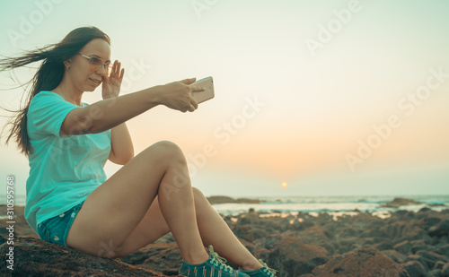 A beautiful young woman is taking selfie on a mobile phone, sitting on the rocks by the sea against the sunset.