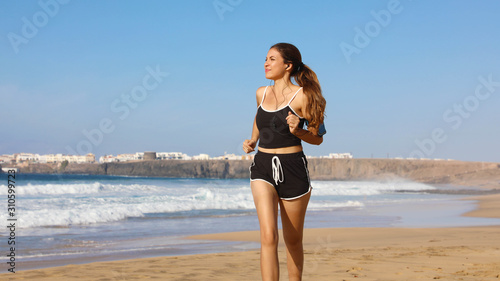 Running woman listening to music on her phone sports armband with touchscreen and earphones on summer beach. Fitness girl jogging training cardio and glutes.