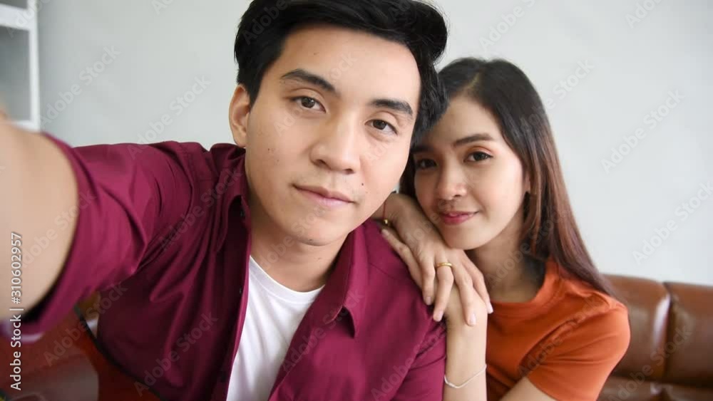 Young Asian couple lover sitting on couch and take a selfie together in modern living room background Young Asian couple lover sitting on couch and take a selfie together in modern living room background
