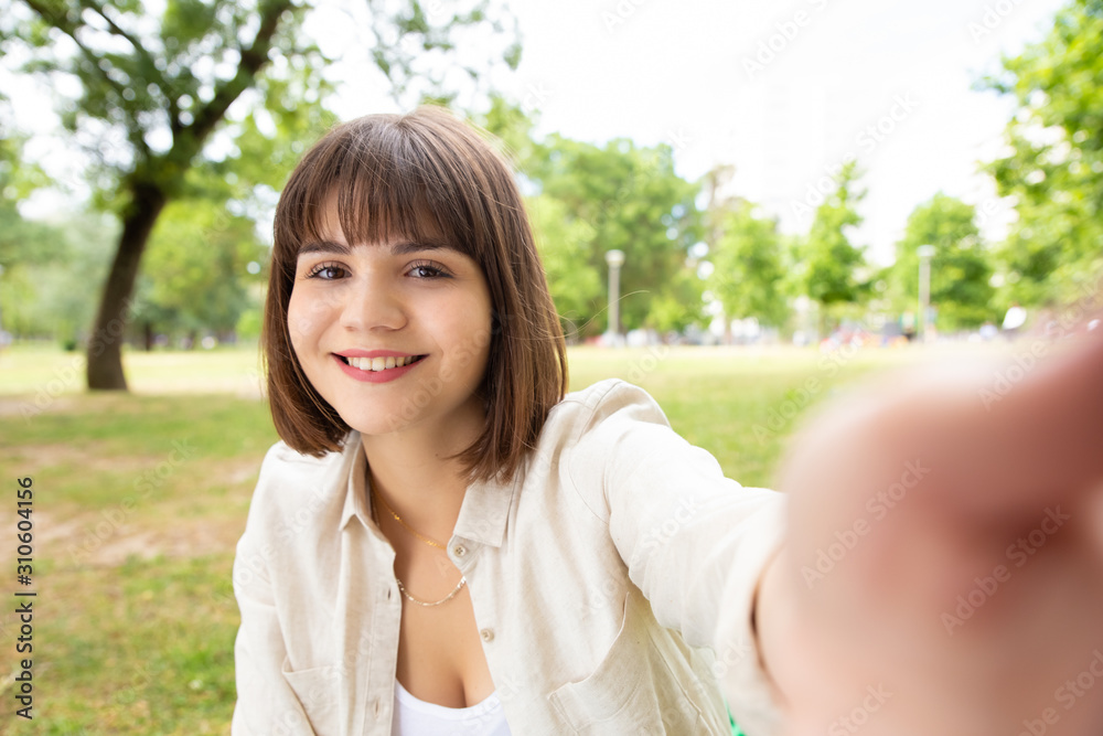 Cheerful young woman taking selfie in park. Beautiful happy young woman smiling at camera in park, selective focus. Facial expression concept