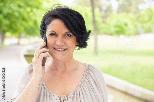 Content woman talking by smartphone outdoors. Beautiful middle aged woman talking by cell phone and smiling at camera in park. Communication concept
