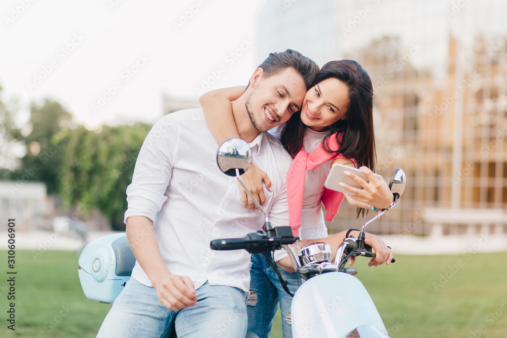 Elegant girl gently touching handsome man on scooter and making selfie with him. Amazing brunette female model taking picture of herself on outdoor date.