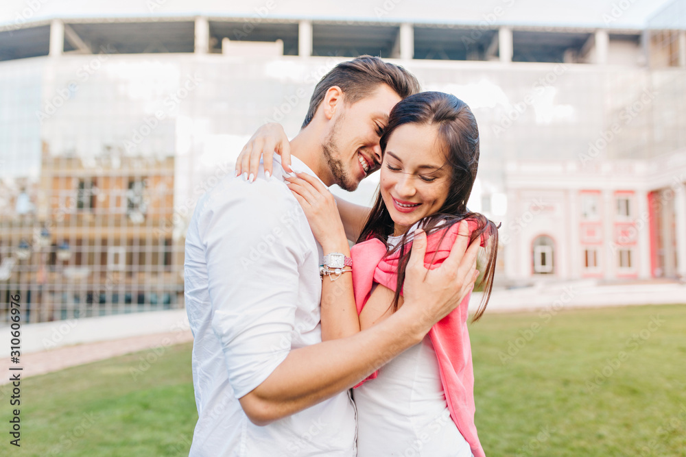 Naklejka premium Smiling girl wears trendy accessories embracing boyfriend during photoshoot on urban background. Handsome man in white shirt enjoying date with joyful brunette girl with pink sweater.
