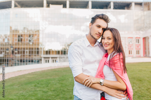 Young married couple posing together in front of modern building during joint weekend. Gorgeous dark-haired european girl huggs with boyfriend on outdoor photoshoot.