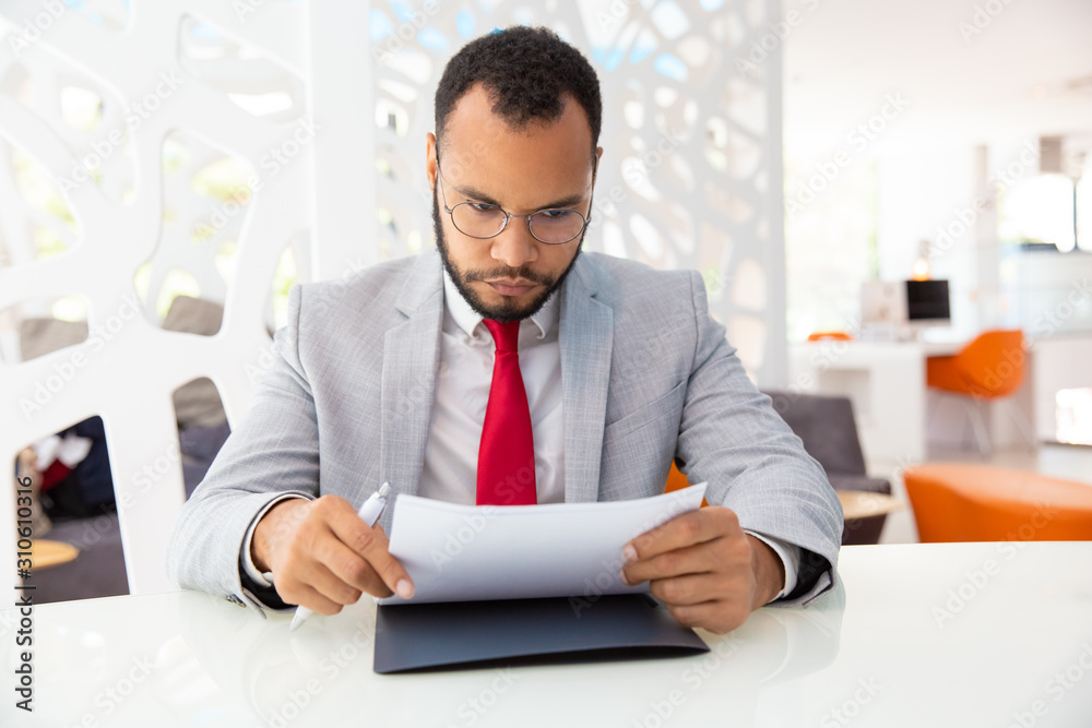 Serious businessman working with papers. Focused young African American businessman in eyeglasses sitting at table and holding documents in office. Paperwork concept