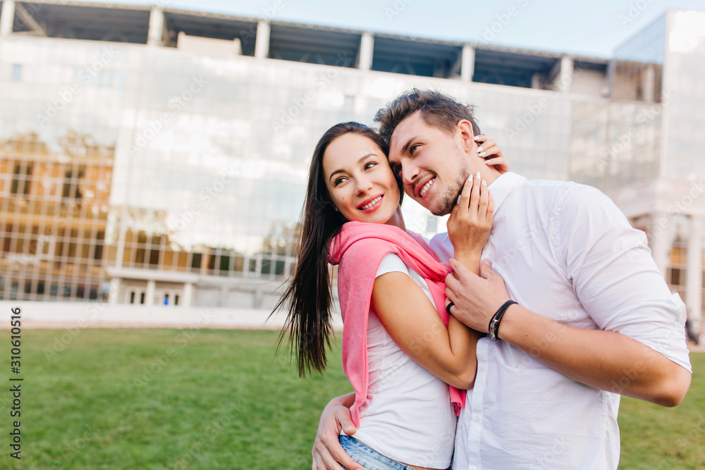 Sporty man wears black bracelet enjoying free time with charming girl and smiling. Amazing dark-haired woman embracing with husband on outdoor photoshoot in city.