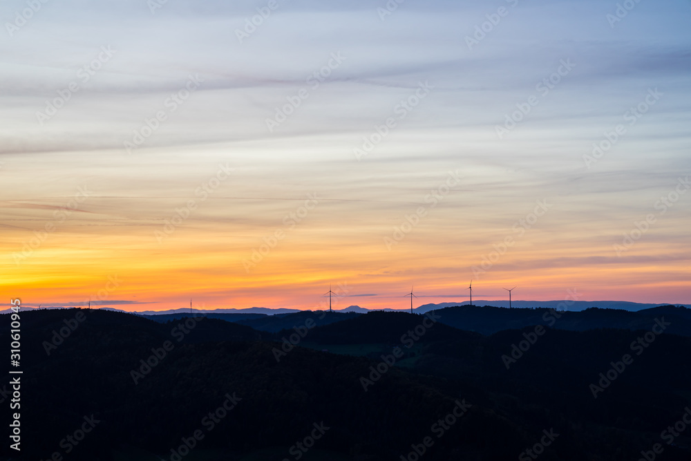 Germany, Orange sunset sky over silhouette of many wind engines in mountainous black forest nature landscape as a symbol for future energy production