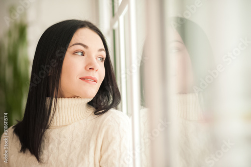 Portrait of a brunette girl at the window with reflection closeup. Young woman in a white knitted dress on a background of a large window and copy space. Free Download Portrait of a brunette girl at the window with reflection closeup. Young woman in a white knitted dress on a background of a large window and copy space. - GFXLoad