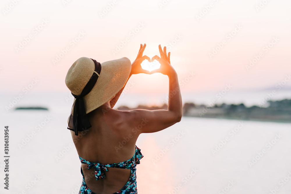 Young woman making a heart shape into the sunset by the sea in sunset
