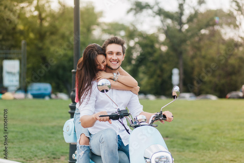 Excited girl in bracelets huggs boyfriend while he driving down the park alley. Outdoor photo of lovely brunette lady enjoying weekend with husband on scooter. photo