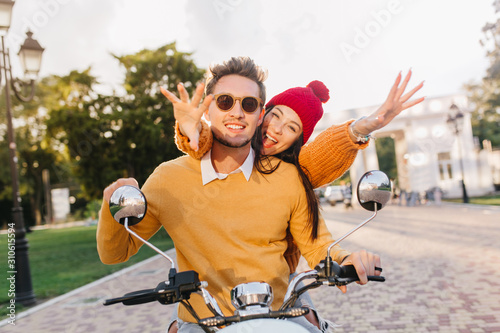 Lovely girl in cute red hat waving hands enjoying extreme date with boyfriend. Handsome man in dark glasses driving scooter in weekend with girlfriend.