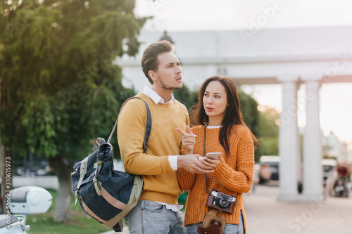 Travelling couple looking around with worried face expressions. Disappointed caucasian man with backpack holding hands with serious girl in knitted sweater with camera.