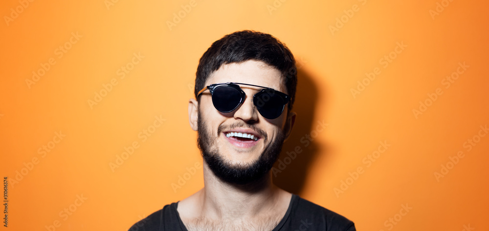 Fototapeta premium Close-up portrait of young modern man laughing, wearing sunglasses on yellow background with copy space.