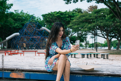 One Asian female in floral dress holding mobile phone in an outdoor park.