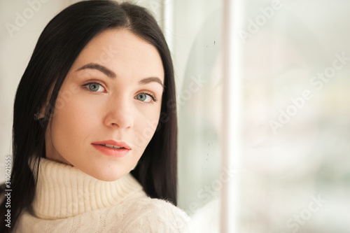 Portrait of a brunette girl at the window with reflection closeup. Young woman in a white knitted dress on a background of a large window and copy space.