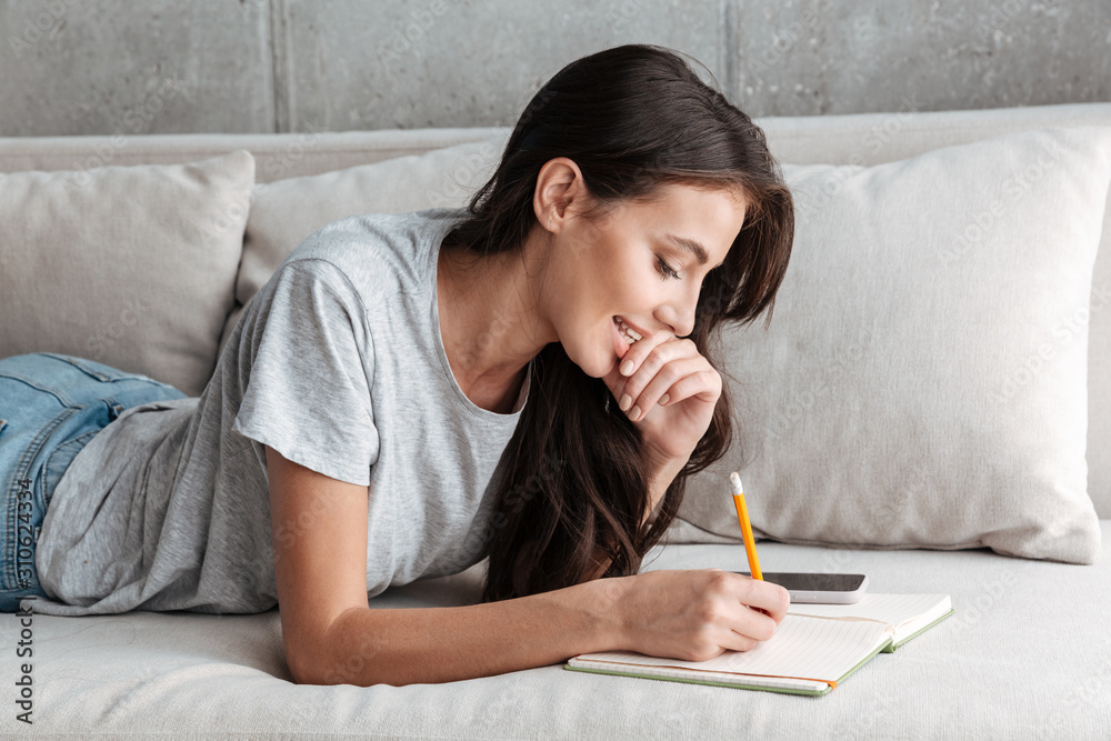 Image of young woman writing down notes in diary book on sofa at home Image of young woman writing down notes in diary book on sofa at home