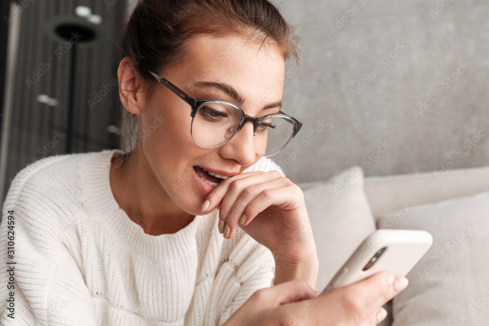 Image of woman in eyeglasses smiling and holding cellphone at home Image of woman in eyeglasses smiling and holding cellphone at home