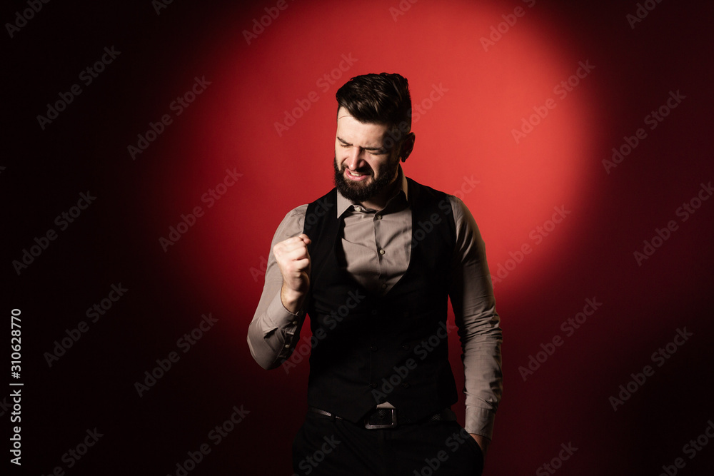 Portrait of a young serious bearded man in a black vest on red background. The man clenches his fist in delight and shouts with joy. Copy space Portrait of a young serious bearded man in a black vest on red background. The man clenches his fist in delight and shouts with joy. Copy space