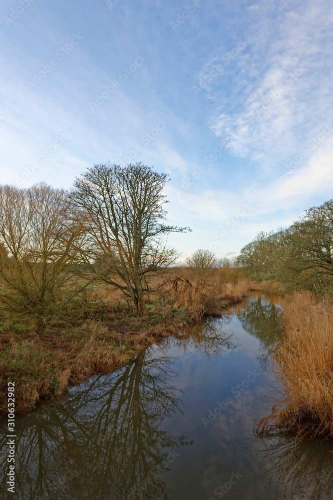 The River Lunan gently running south amongst the Fields and countryside with the reflections of the riverbank trees in its waters.