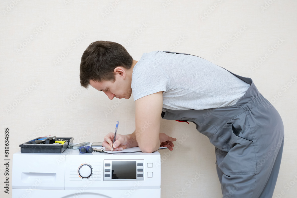 young serviceman in grey uniform with set of tools writes on clipboard examining modern washing machine with screen on control panel in room