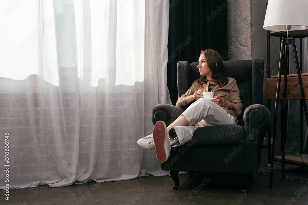 Obraz premium Pensive girl with prosthetic leg holding coffee cup and looking at window in living room