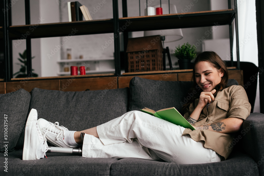 Obraz premium Smiling woman with leg prosthesis reading book on couch in living room