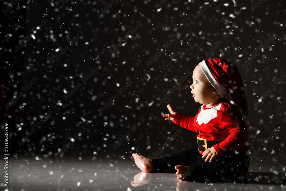 Infant baby boy toddler in red christmas cap and new year costume is sitting on ice under the snow looking at light ray Infant baby boy toddler in red christmas cap and new year costume is sitting on ice under the snow looking at light ray