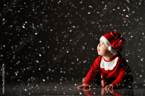 Infant baby boy toddler in red christmas cap and new year costume is crawling on all fours under the snow looking up