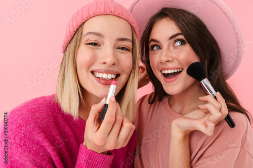 Image of two joyful women smiling and applying makeup with cosmetics photo