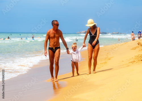 family with little dother holding hands and walking on sand tropical beach waves and people in the background
