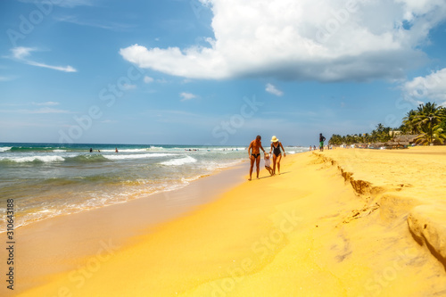 young tanned family walk hand in hand with little daughter on sandy shore of tropical ocean beach, Sri Lanka.