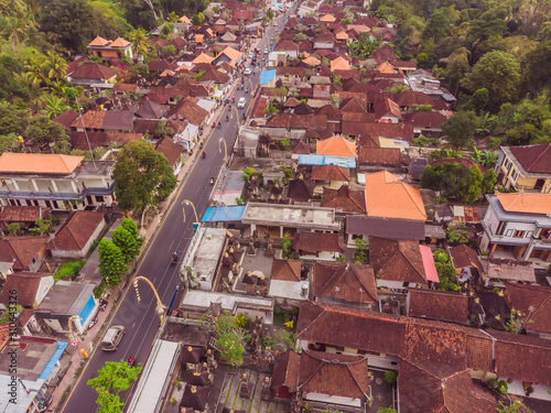 Many villas with brown-orange shingle roofs between tropical trees on the sky background in Ubud on Bali. Sun is shining onto them. Aerial horizontal photo