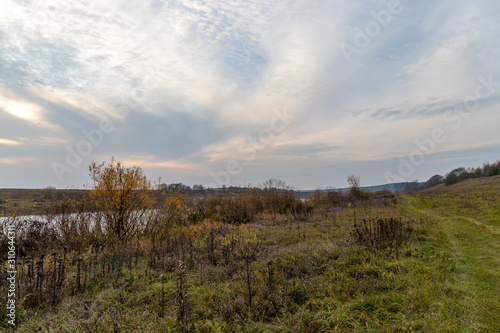 Autumn landscape. A vague spot of the setting sun in the sky with wavy stripes of clouds. Valley of the river, overgrown with withering grass and bushes. A curved dirt road runs along the shore
