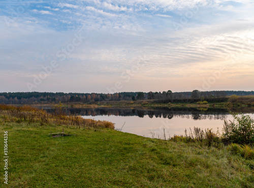 Calm landscape at eve dusk. Green bank with a place for relaxation and a picnic with beautiful view of the river  distant forest. The still mirror of the water reflects trees and sunset in cloudy sky
