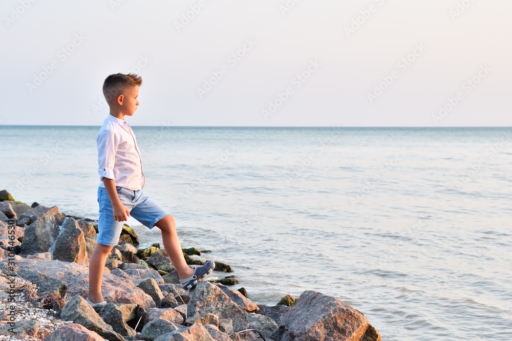 Cute boy standing on the stones on the seashore watching the sunset