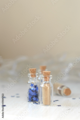 Little glass bottles with a brown letter and blue stars are on the white wooden table lights background bokeh. Concept of secret message in a bottle, keep dreams. Magic background.