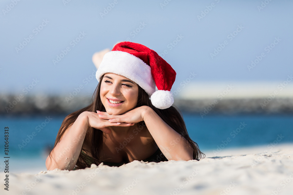 Obraz premium Portrait of happy young woman on tropical beach wearing Santa hat enjoying winter holidays in exotic destination. Bikini girl sunbathing on white sand beach loving vacations