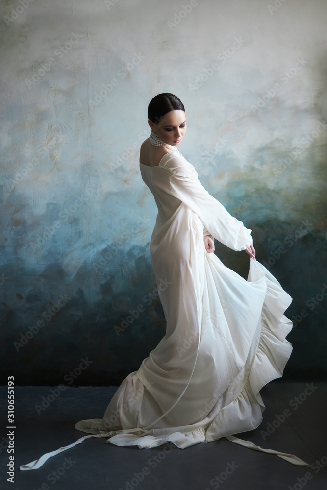 Beautiful slender brunette girl sitting on the sofa in a long white dress. Portrait of a woman with a jewellery on neck. Perfect hairstyle and cosmetics of the woman, new collection of light dresses