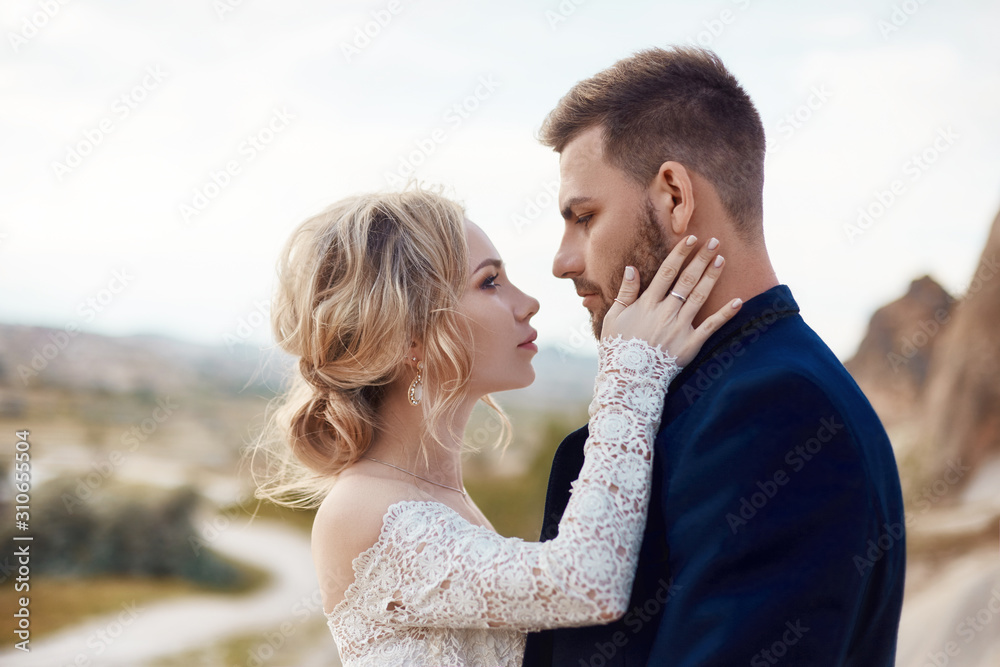 Couple in love hugs and kisses in fabulous mountains in nature. Girl in long white dress with bouquet of flowers in her hands, man in jacket. Wedding in nature, relationships and love