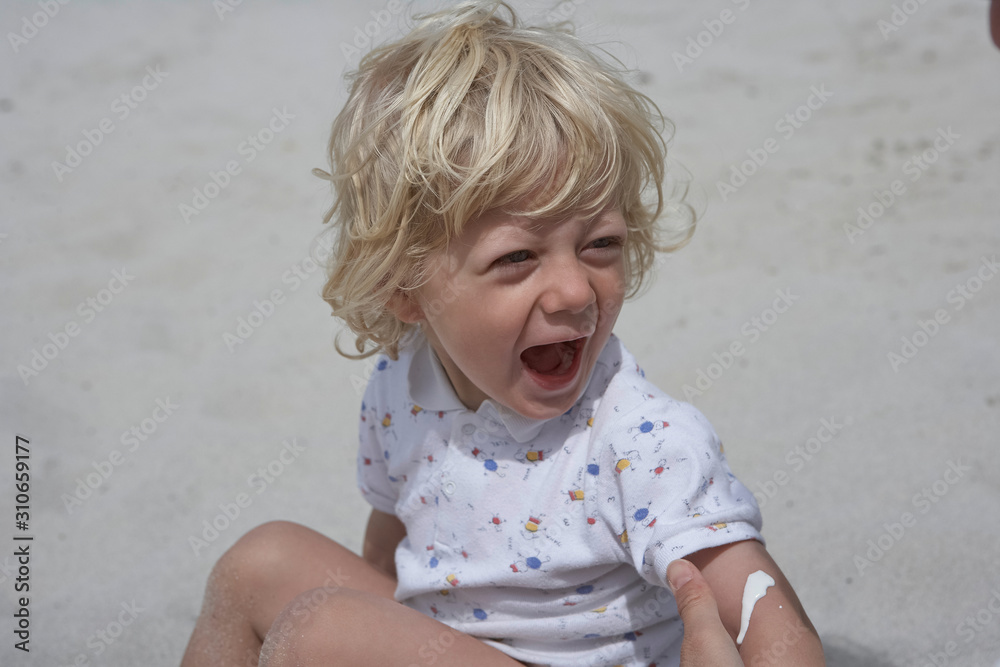 Cute little boy happily laughing sitting on a sandy beach while his arm is moisturized Cute little boy happily laughing sitting on a sandy beach while his arm is moisturized