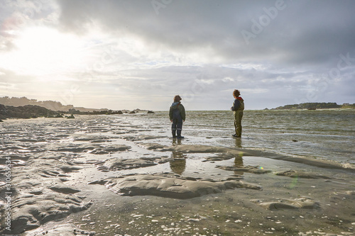 Two boy friends in rainwear standing on the sand of a beach in Brittany at low tide, looking into the sea, wind in their hair, a small island in the background, the sun begins to shine after a rain