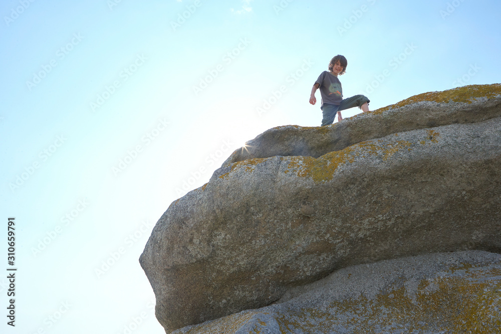Proud boy standing on a big rock, blue sky, sun flares