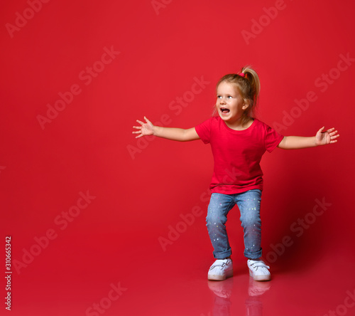 Small happy excited blond girl in stylish casual clothing and white sneakers jumping and feeling playful over red wall background