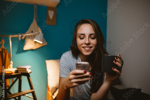 woman in her bedroom relaxing using mobile phone and drinking coffee or tea