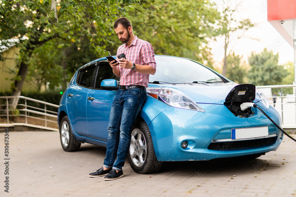 Handsome man is holding a phone while electric car charged at the charging station Handsome man is holding a phone while electric car charged at the charging station