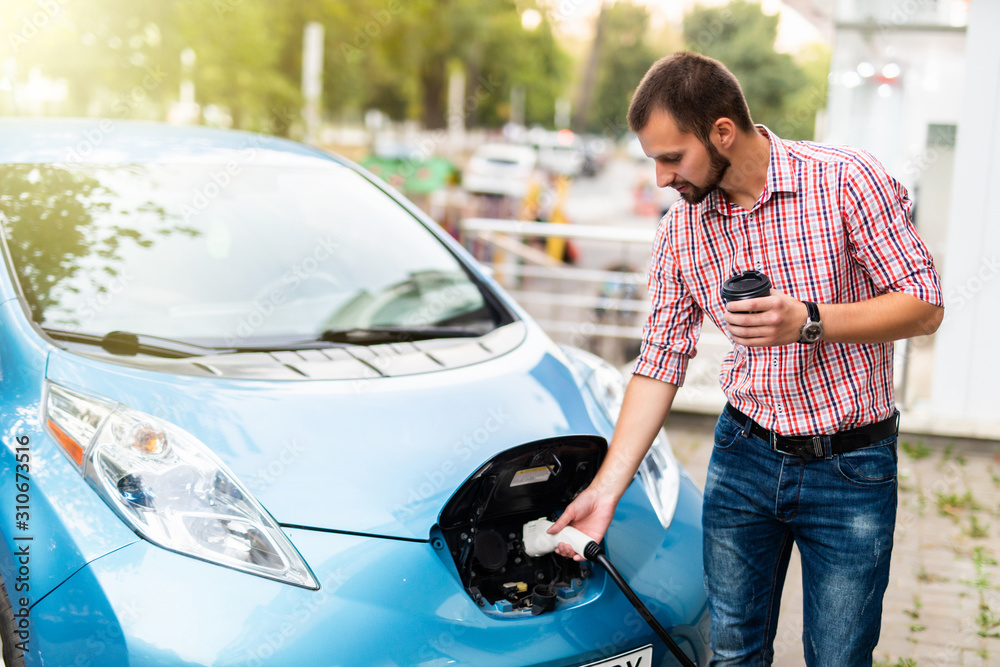 Young man charging his electric car and drinking coffee