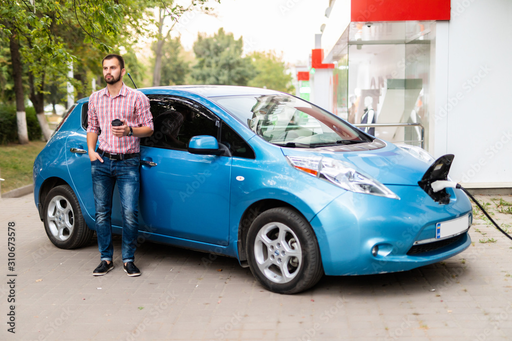 Young handsome man with coffee near his electric car charging at station Young handsome man with coffee near his electric car charging at station