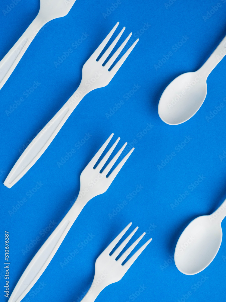 Plastic white disposable fork and spoon on a trendy blue color background. Concept plastic dishes, fast food, plastic pollution. Top view, flat lay.