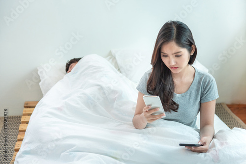 wife spying the phone of her husband while man sleeping in bed at home.asian young girl check and suspension on boyfriend phone while he nap on bed.